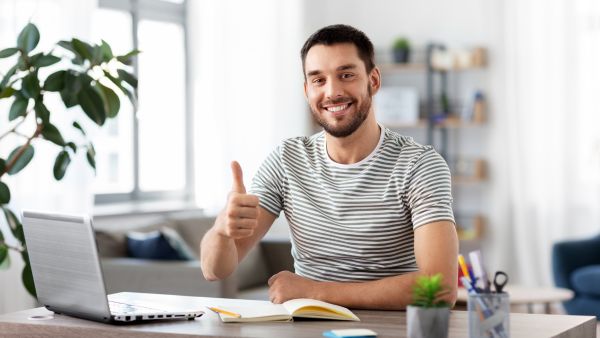 Smiling man at a desk giving a thumbs up with a laptop open beside him, in a bright home office setting.