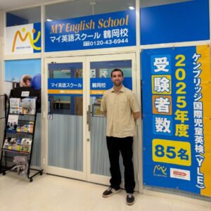 Beige-shirted man stands outside a blue MY English School storefront with a large promotional banner on the right (2025/85名).